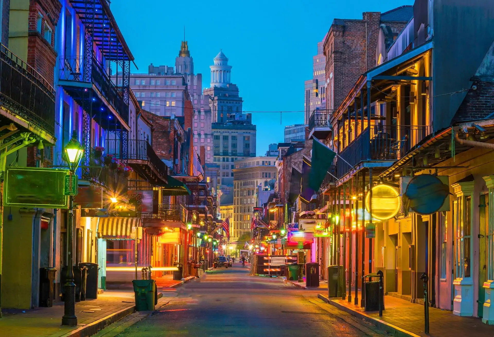 A view of of an empty road in New Orleans with buildings and colorful lights on either side. Historic buildings can be seen in the background.