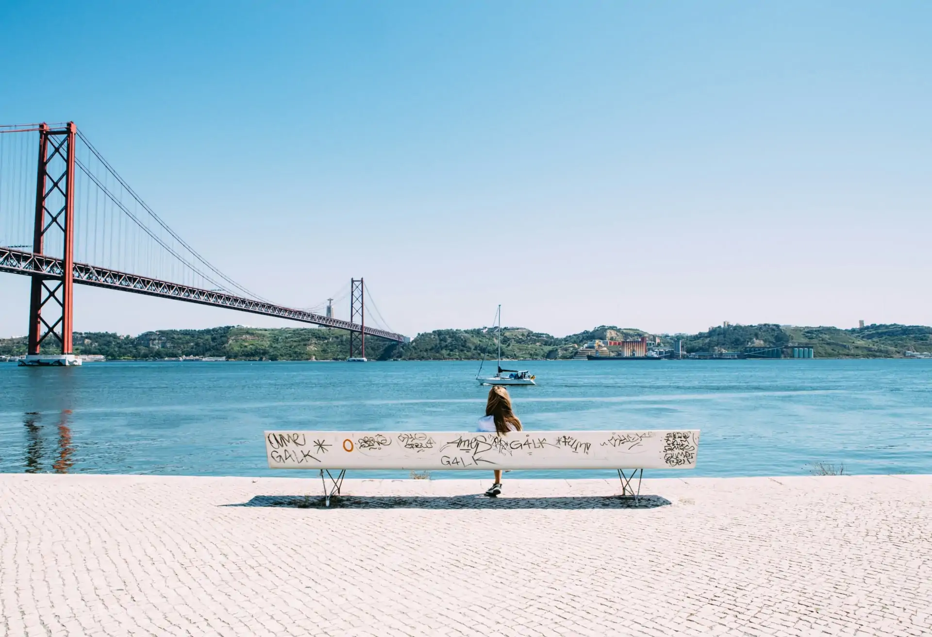 A view of the sea in Lisbon with an industrial bridge and the city in the background. In the foreground, a woman sits on a white bench with graffiti.