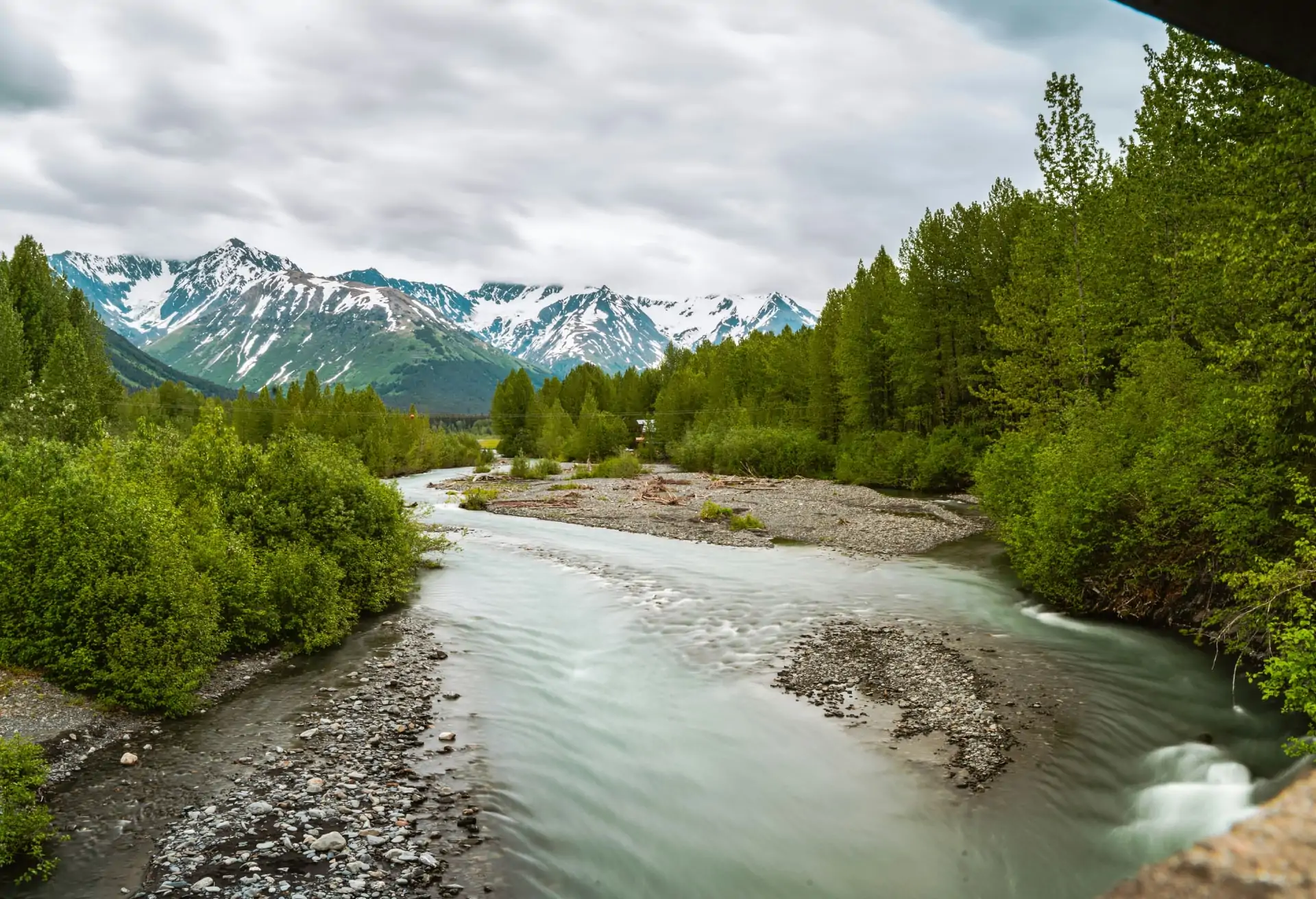 A view of a river, with green trees either side and snow-capped mountains the background.