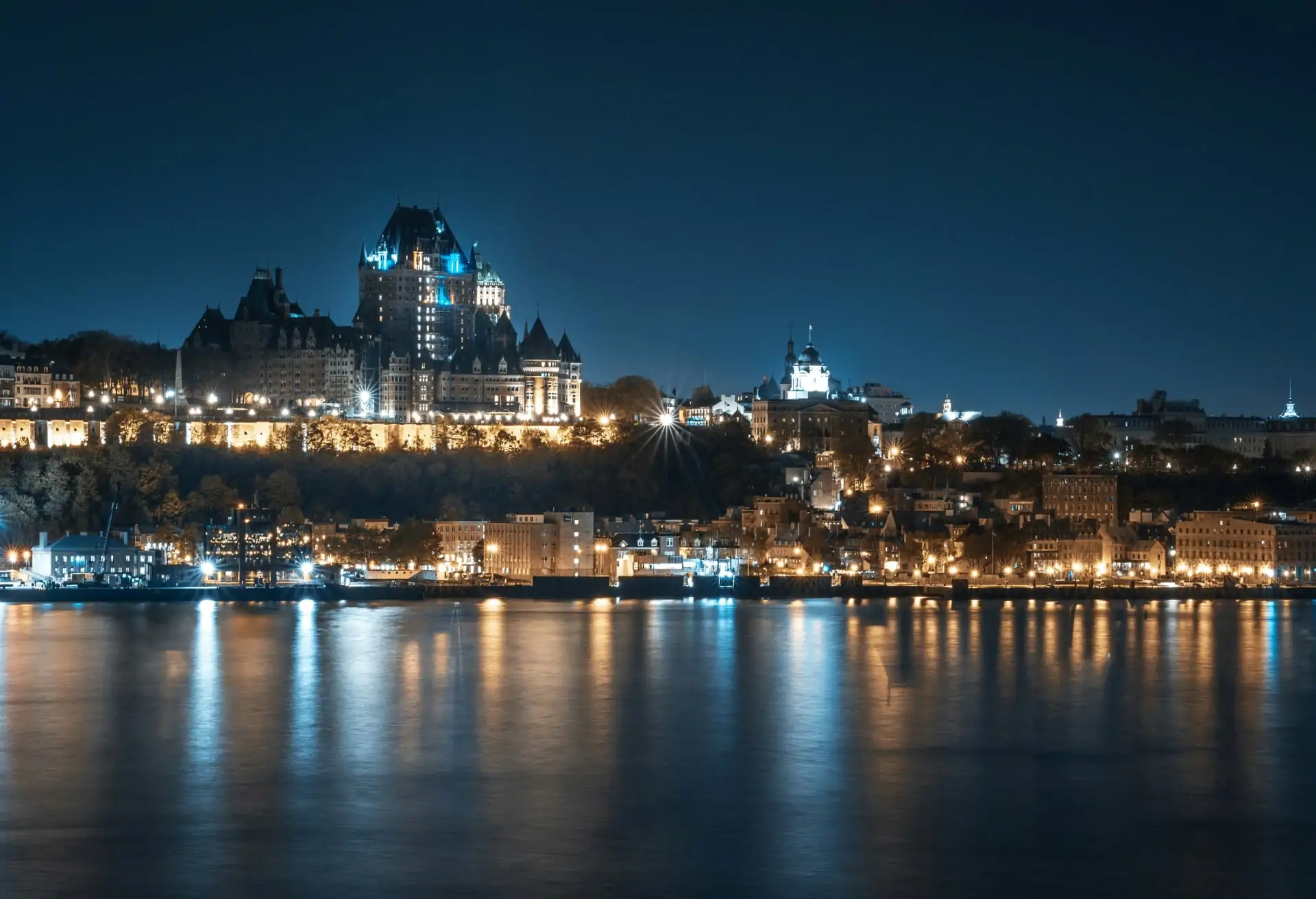 A view of Quebec's old town at night with buildings and lights and the river in the foreground.