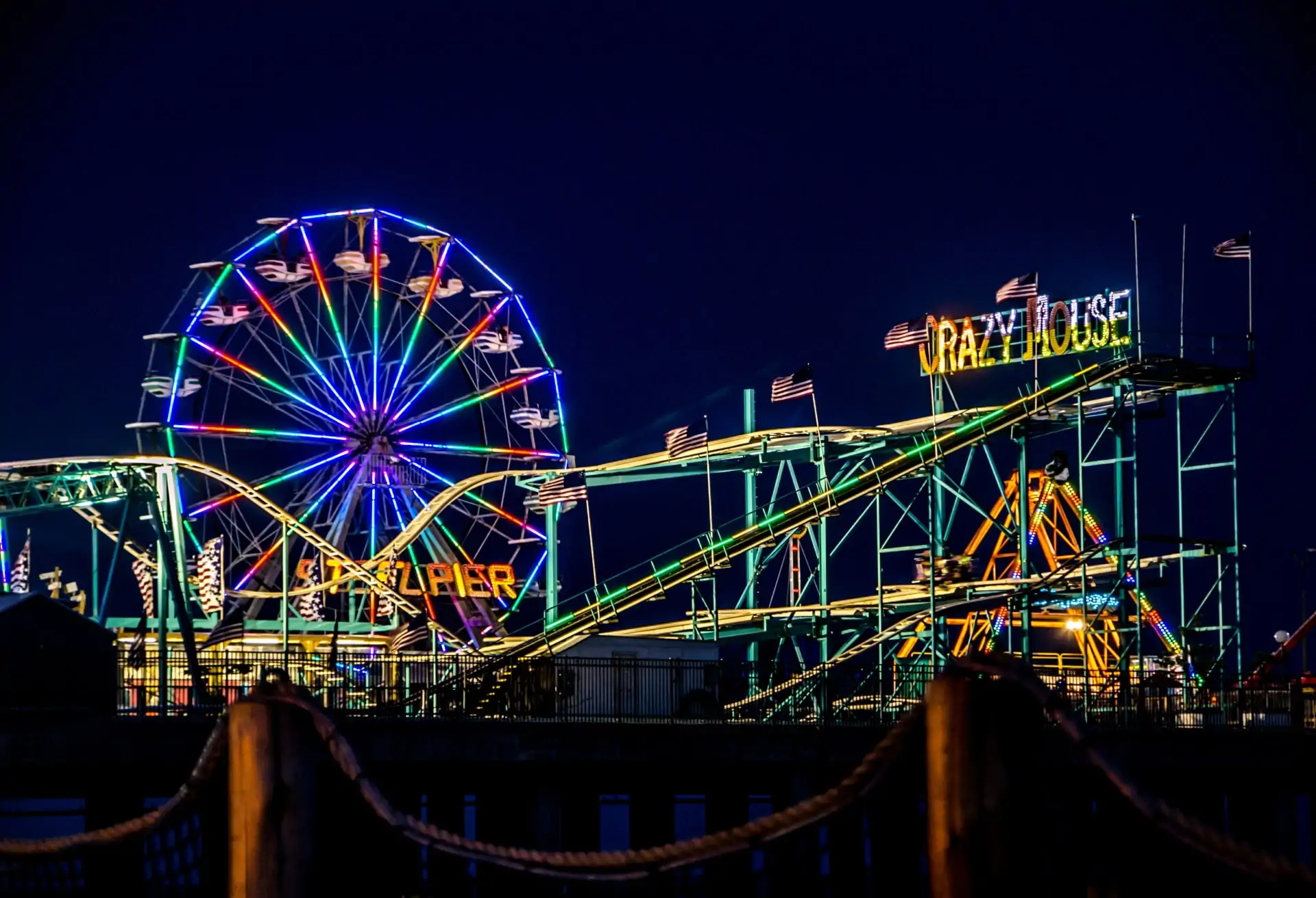 A view of a rollercoast on Atlantic City's boardwalk at night. There's also a Ferris wheel in the background and lots of neon lights.