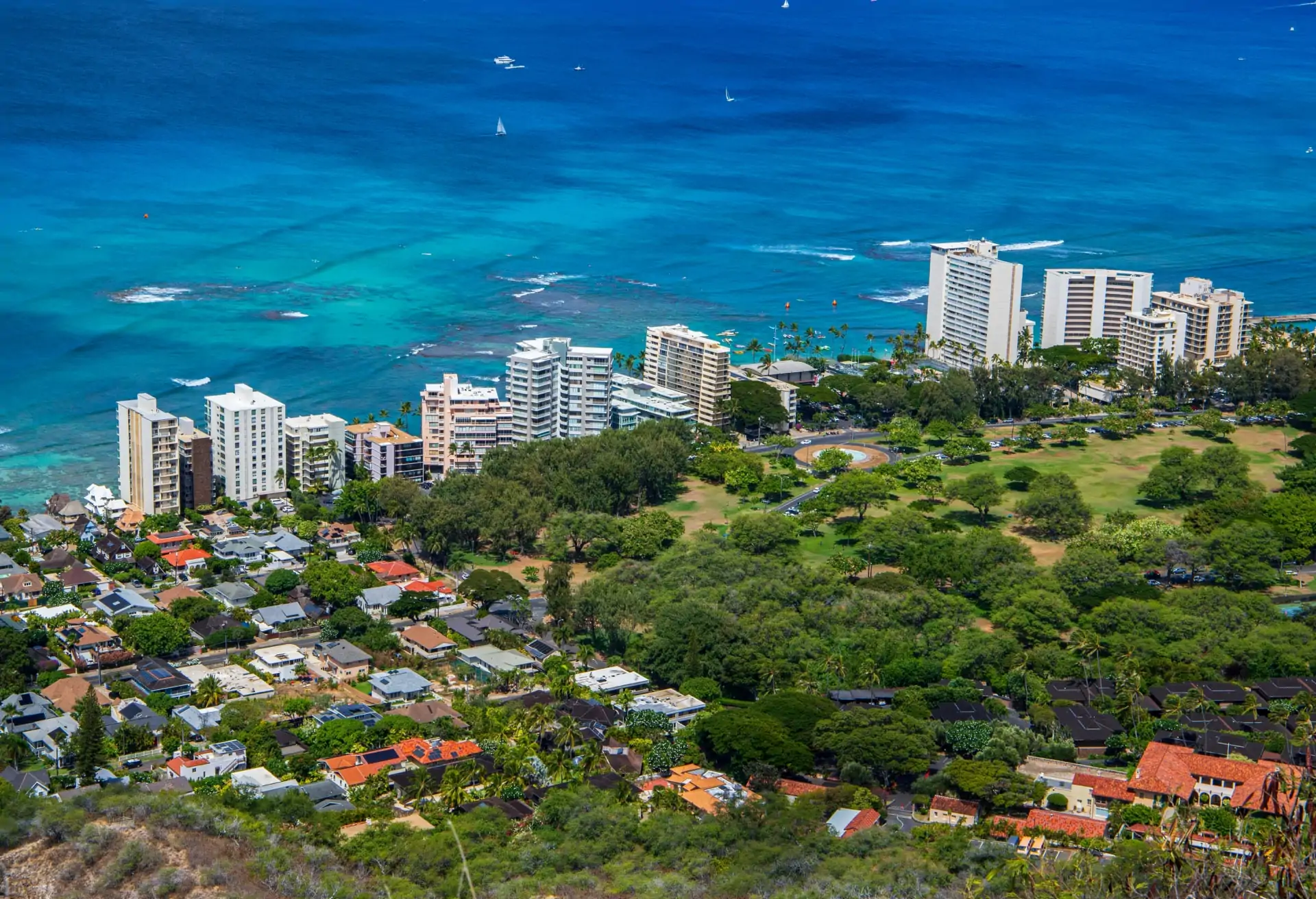 A view of a costal city with modern buildings along the seafront in the background. In the foreground, there are trees and houses.