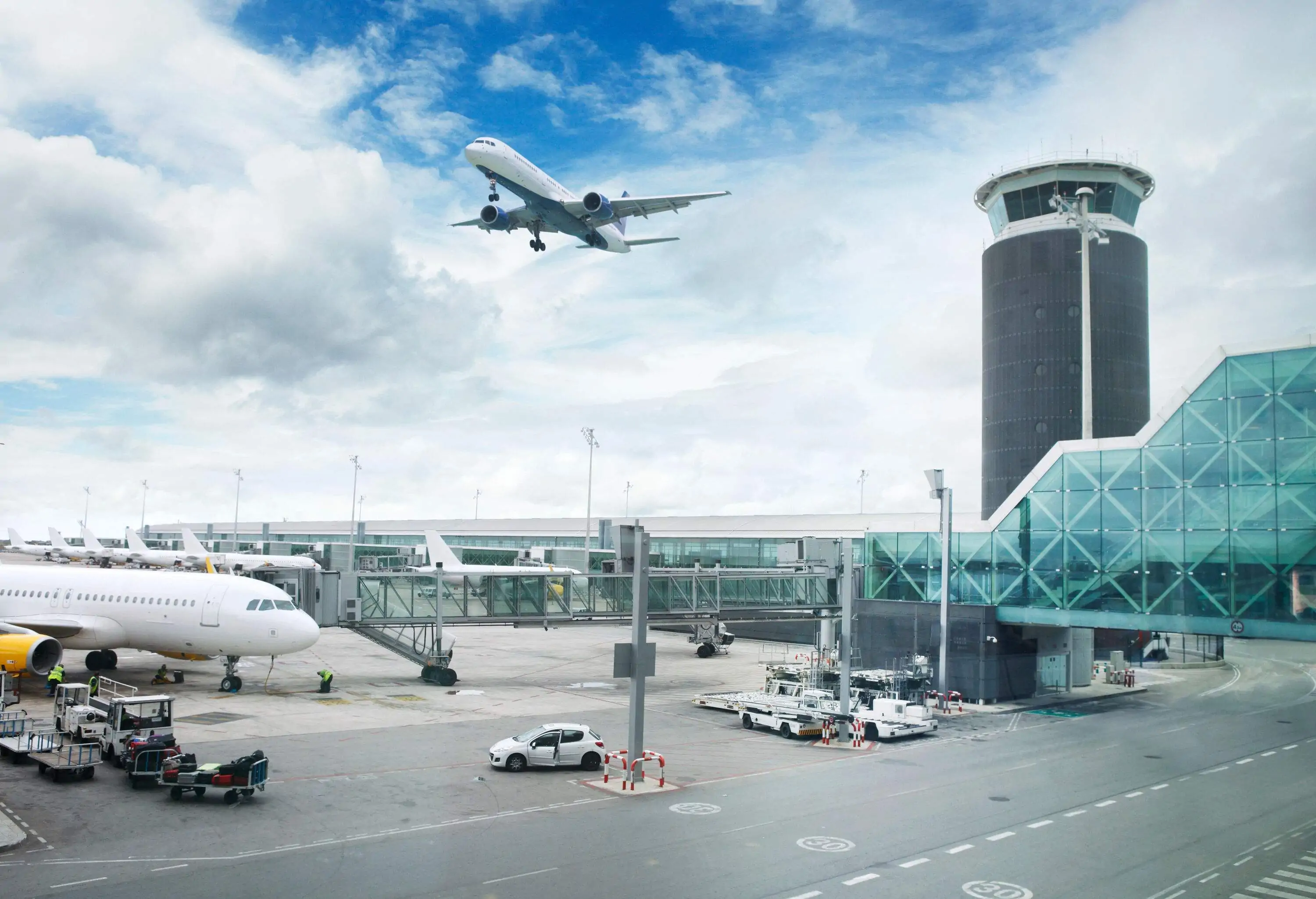 An airplane flying above the airport grounds with the control tower situated next to the jet bridges.