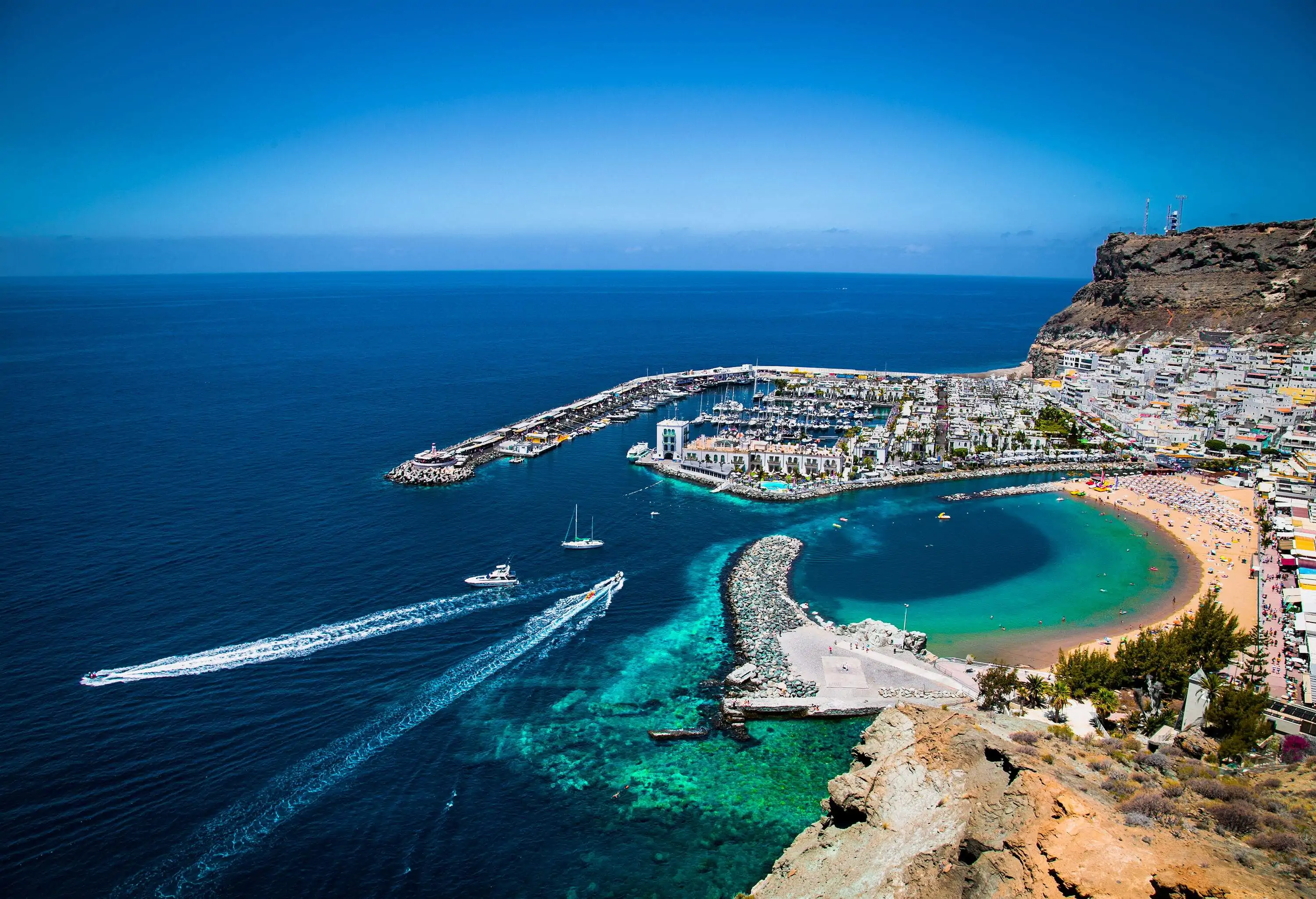 A port town with a packed marina and outdoor cafés along the promenade facing the beach.