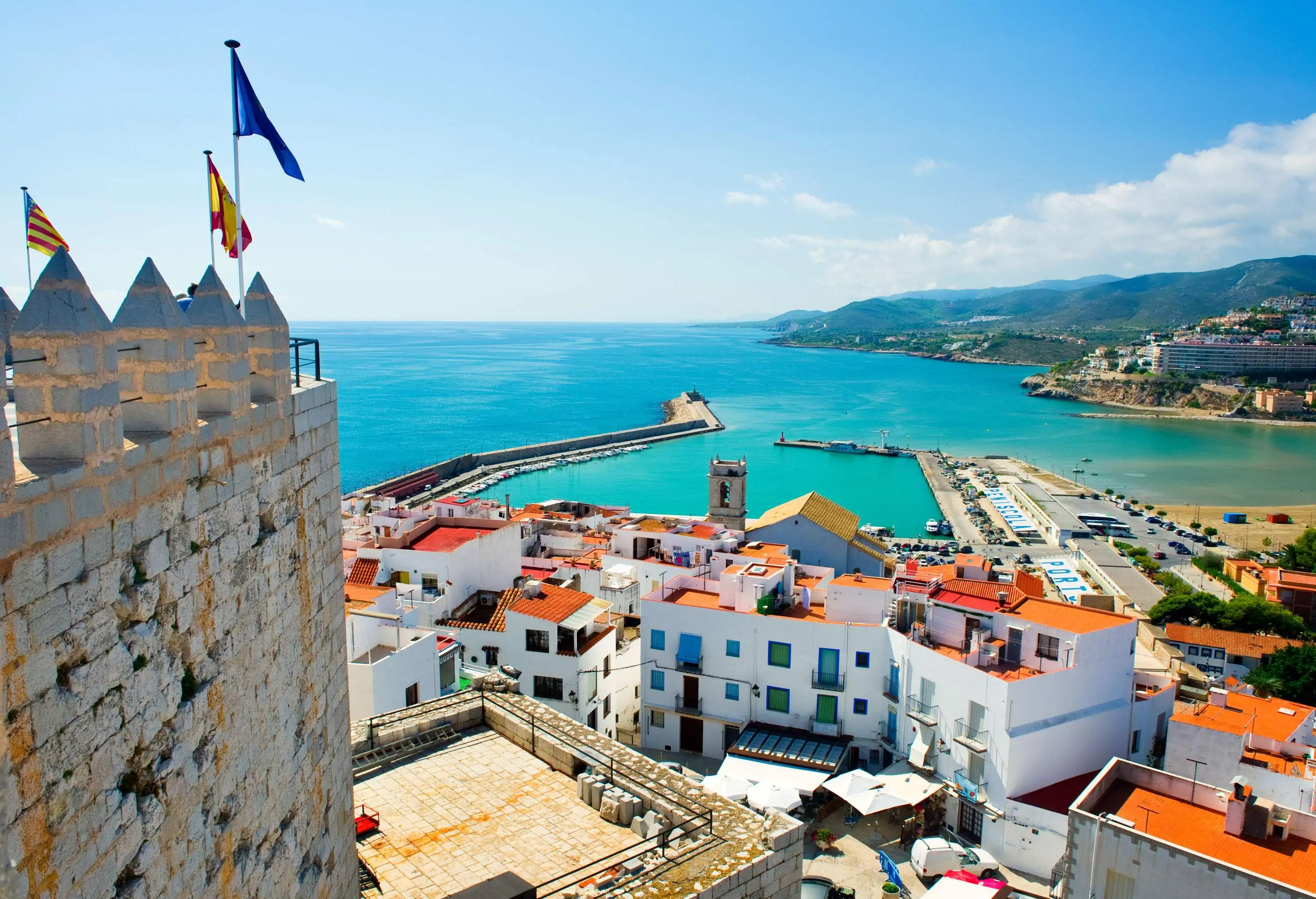 A small port at the foot of a castle along whitewashed houses surrounded by turquoise water.