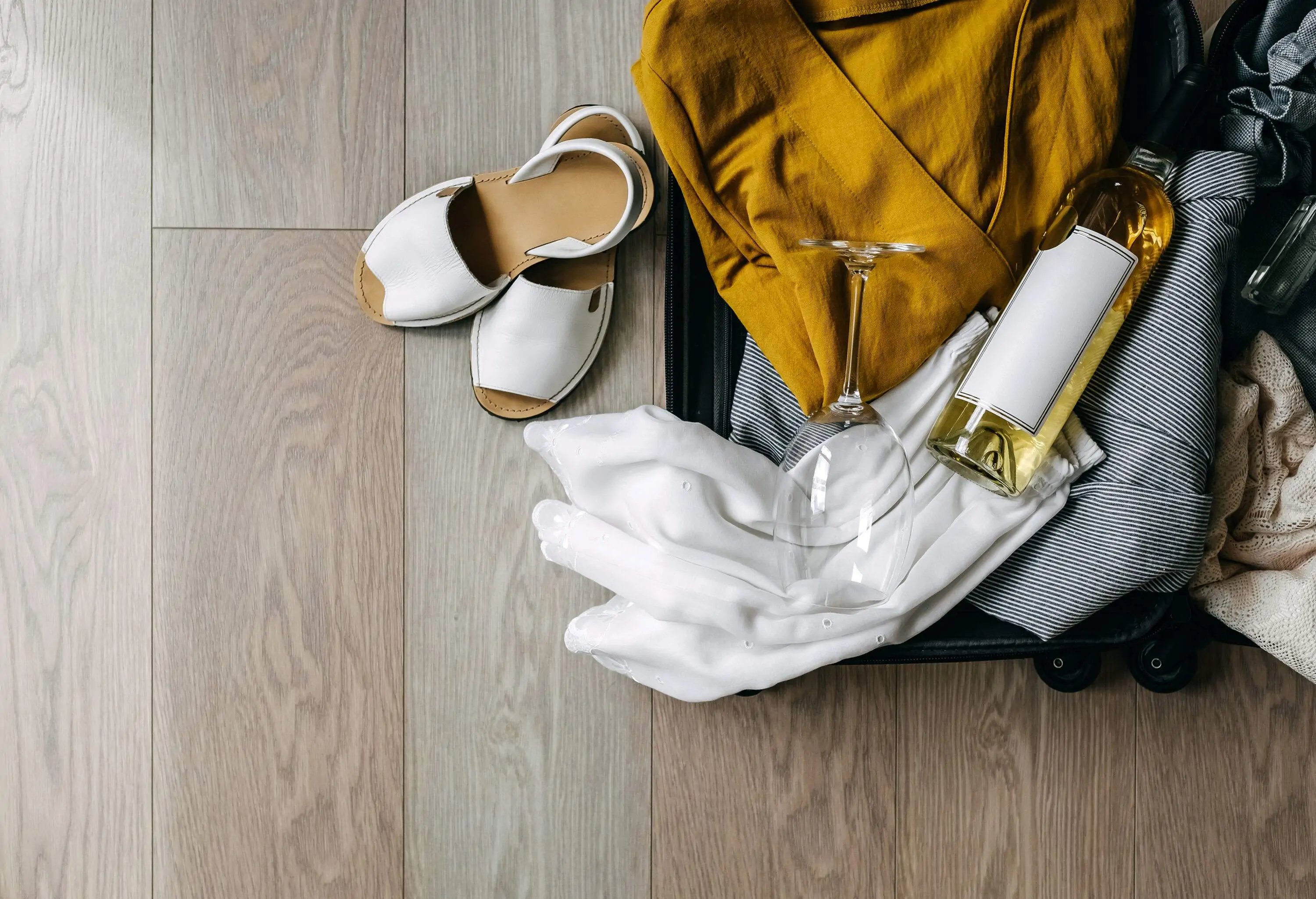 An open suitcase filled with colourful summer clothes, accompanied by a wine glass, a wine bottle, and a pair of sandals.