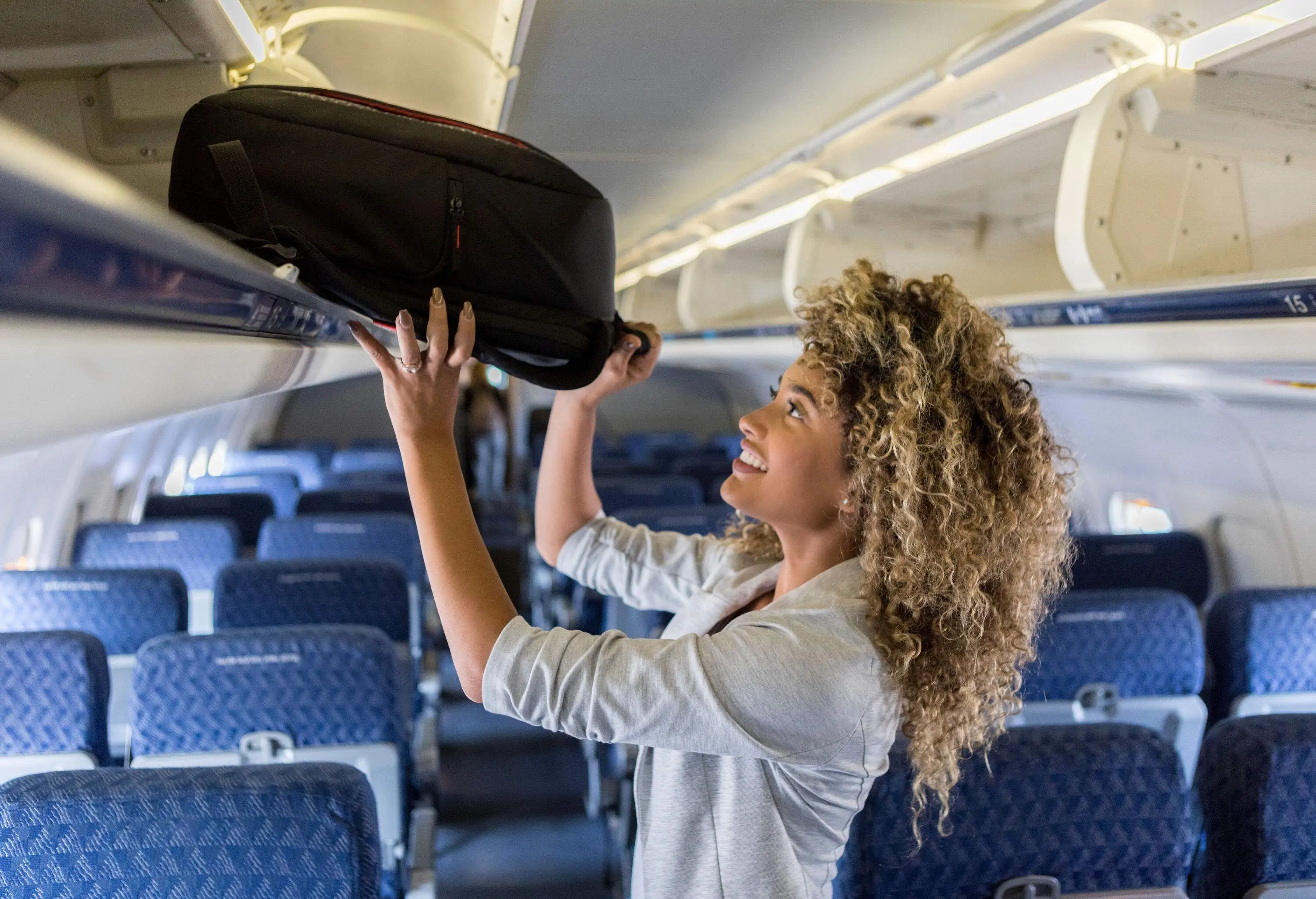 A blonde curly hair passenger smiles while putting her luggage in the overhead bin.