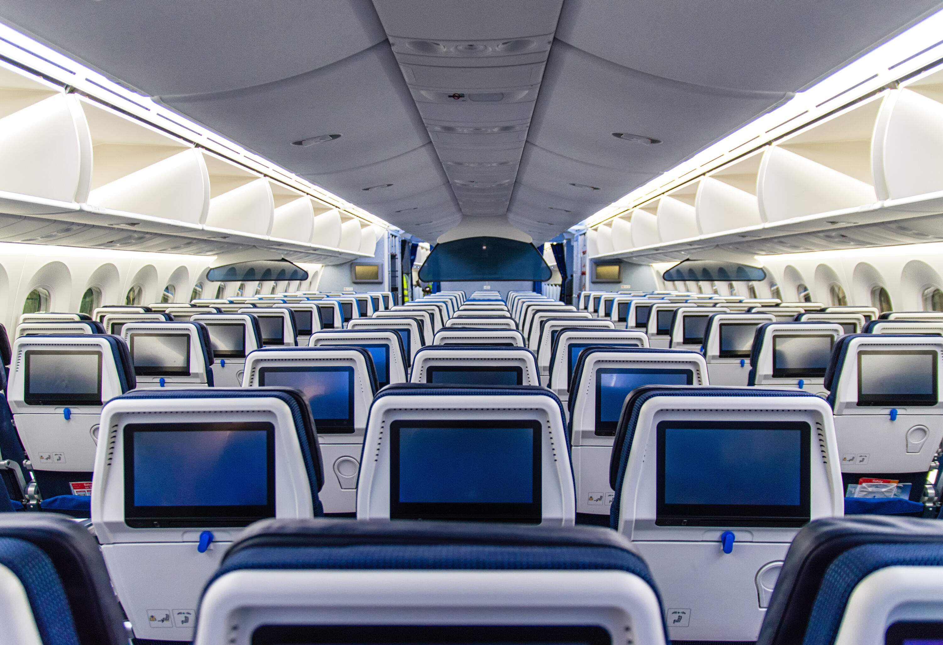 Rows of airplane seats with touchscreen TVs in a cabin.