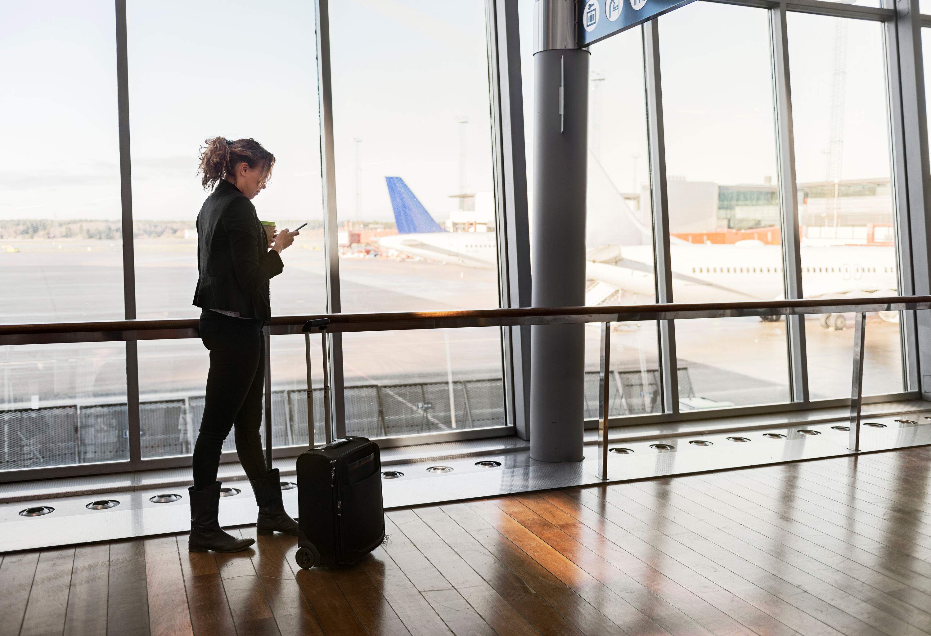 A woman checking her smartphone while standing next to her suitcase inside an airport terminal.