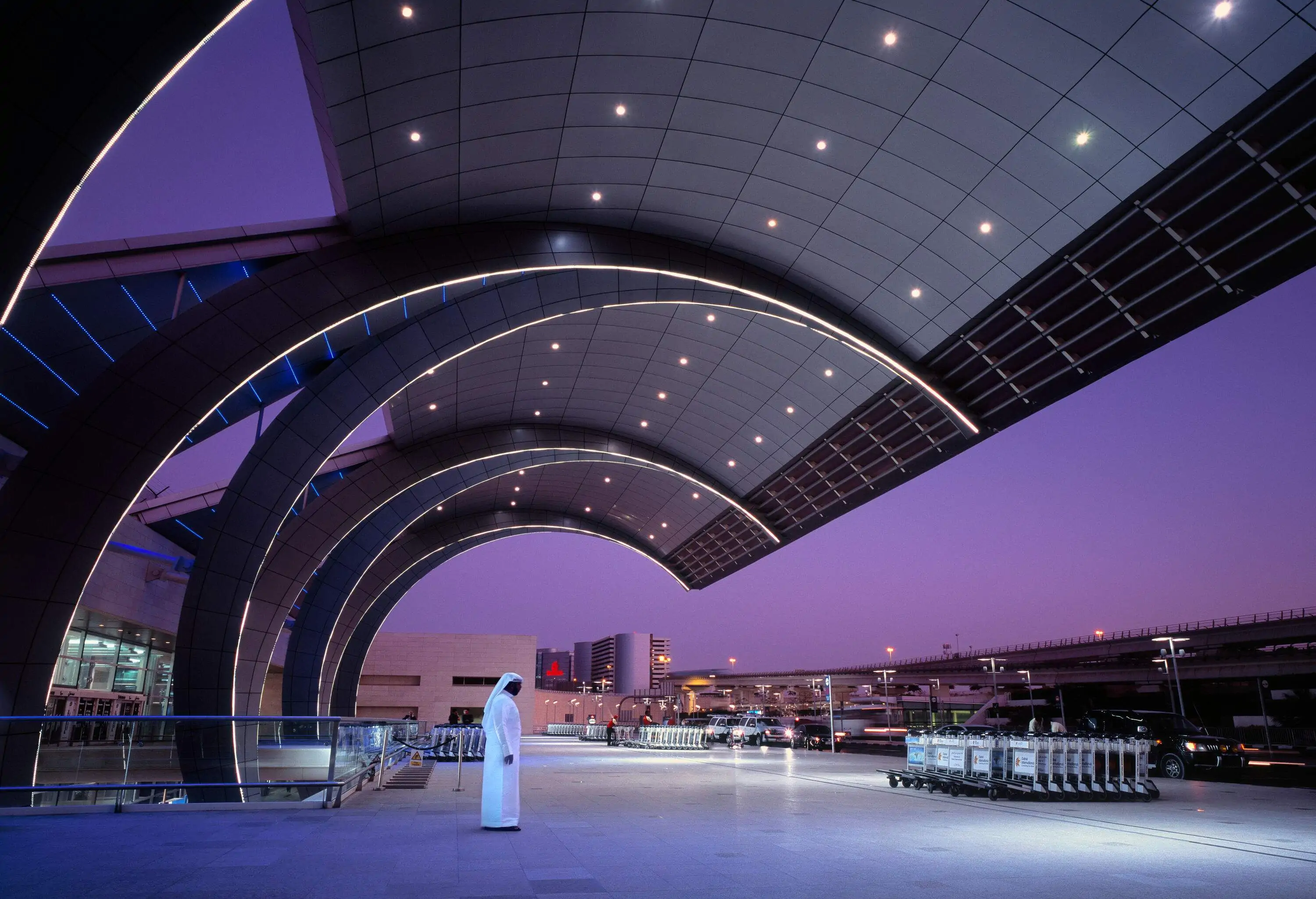 An Arab man in a traditional white dress called Dishdasha stands under the illuminated futuristic curved steel façade of Dubai International Airport Terminal 3 at night.