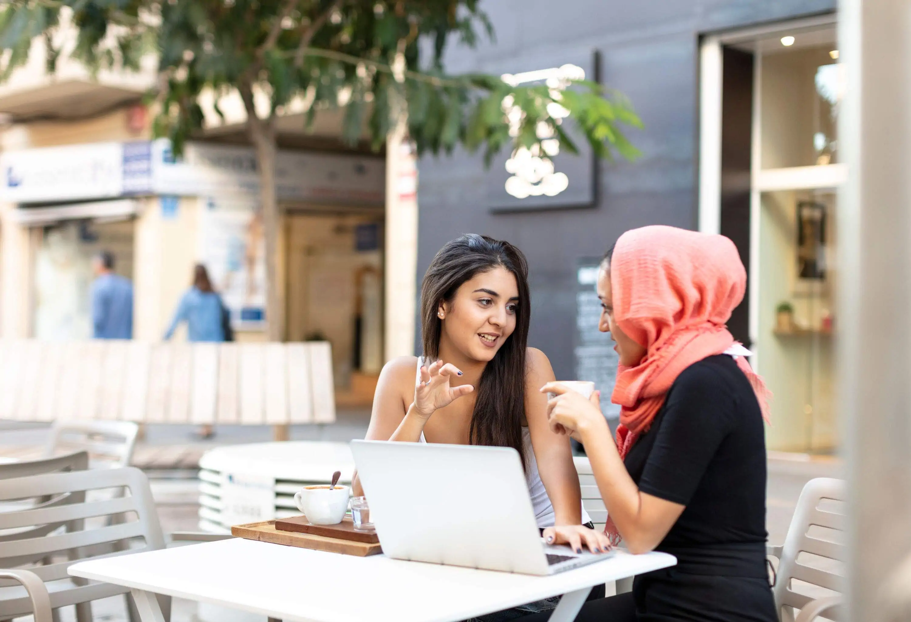 Two friends sit at an outdoor table, engrossed in conversation with a laptop and a cup of coffee resting on the table in front of them, while the background artfully blurs to emphasise their interaction.