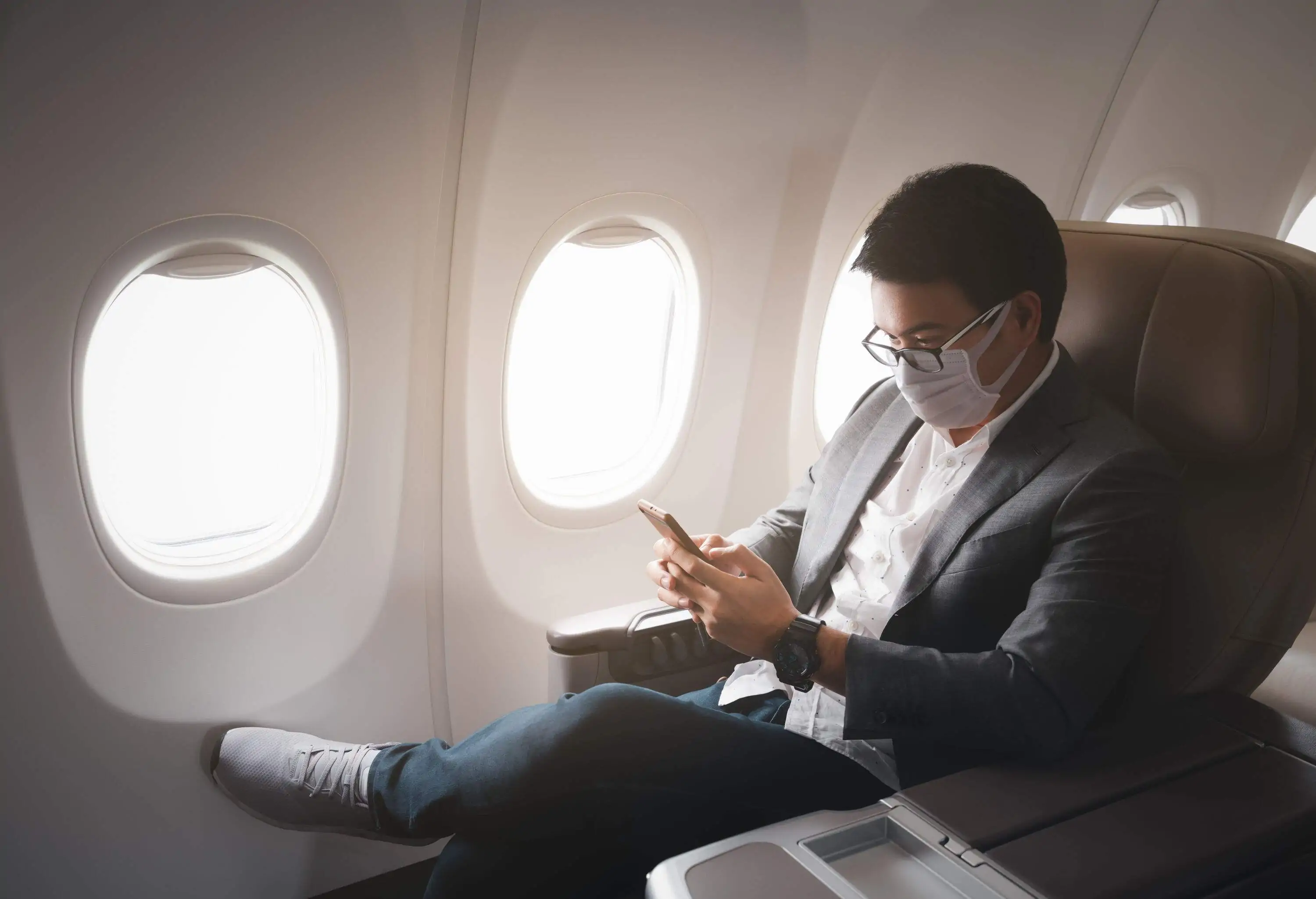 A man in a business casual attire wearing a facemask as he sits through a flight with a smartphone in his hands.