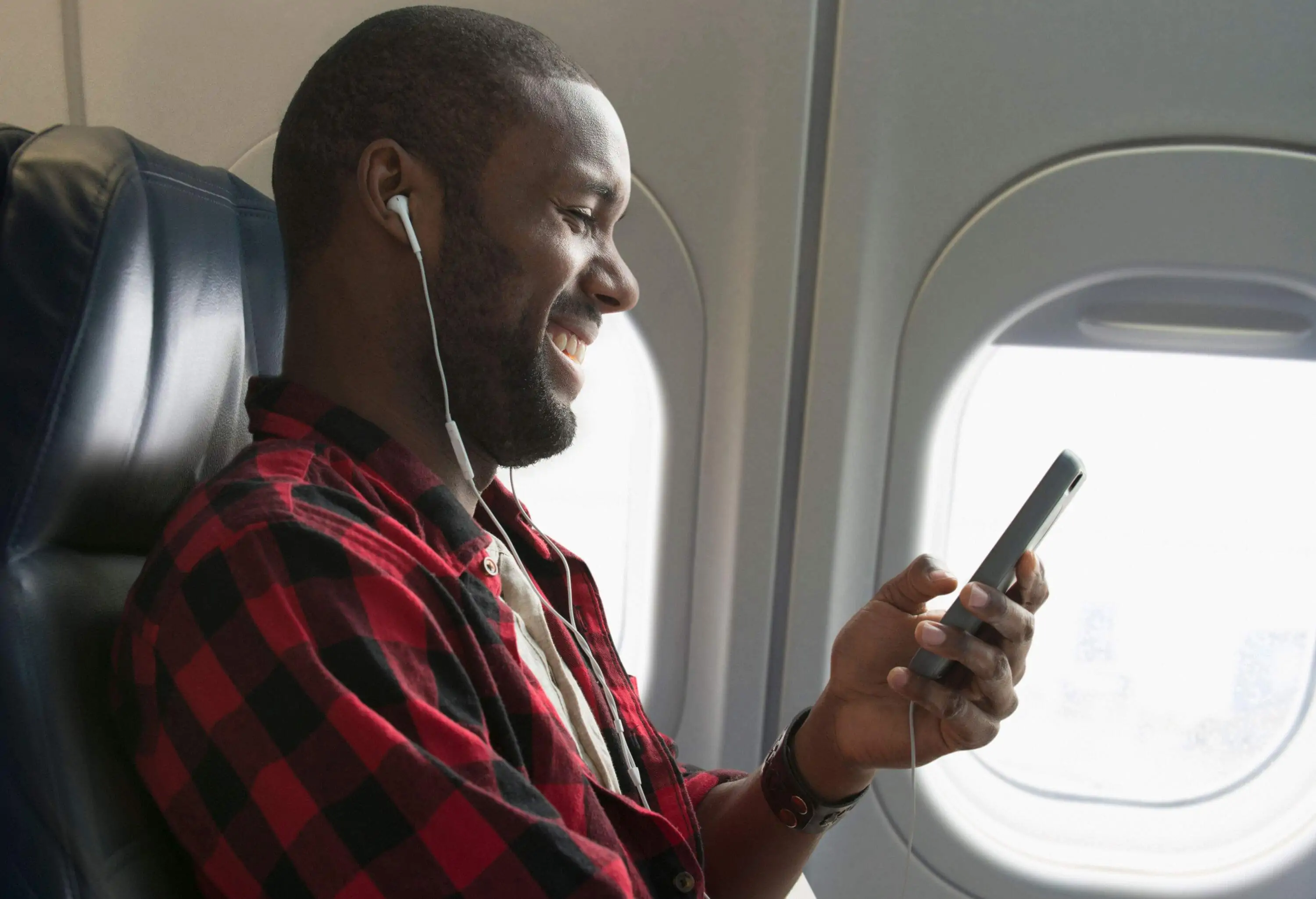 A male passenger with earphones smiling while taking a look at his smartphone on the plane's window seat.